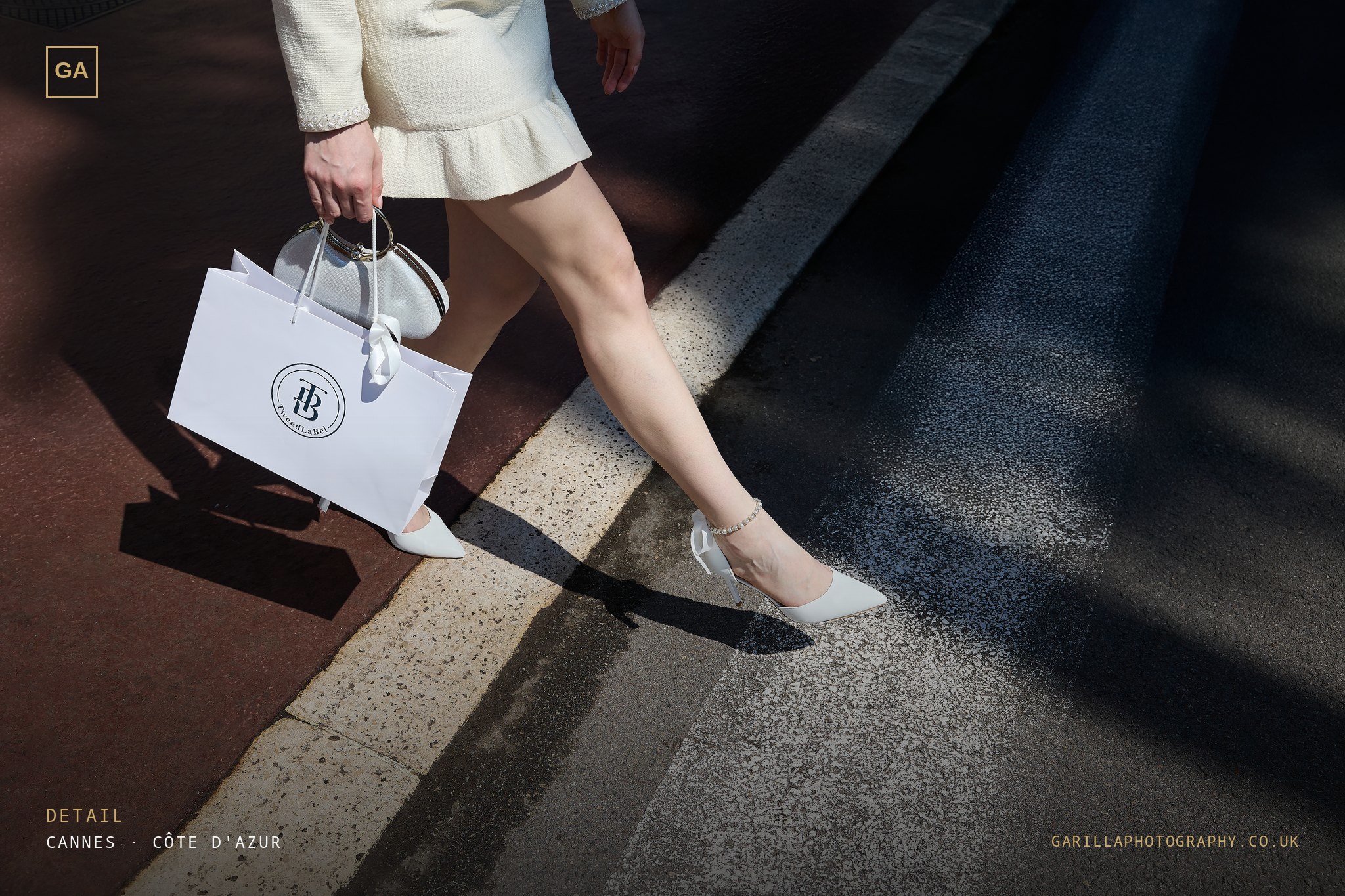 Detail — white heels and the branded shopping bag crossing a sunlit curb